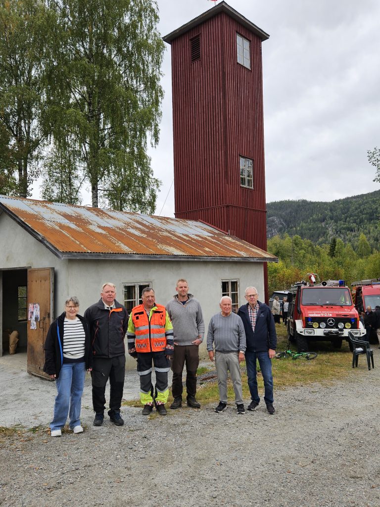 Styret i Venneforeningen gamle begnadalen brannstasjon poserer foran stasjonen med brannbiler i bakgrunnen